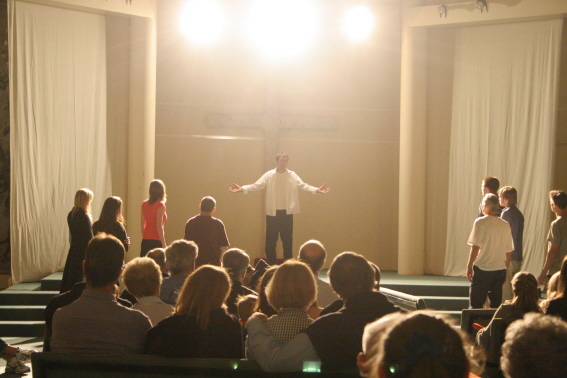 A man stands on the stage, portraying Jesus, under a cross and bright shining lights before a crowded room
